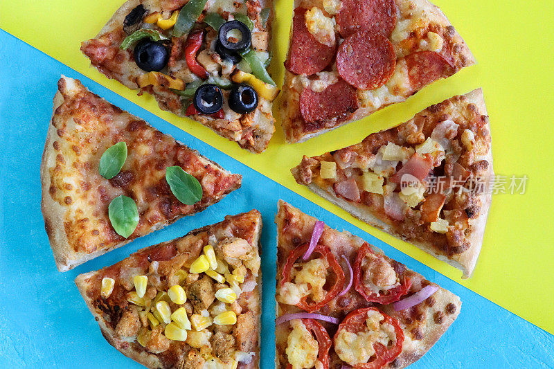近距离图像的披萨片与各种浇头，包括夏威夷披萨，鸡肉和甜玉米，意大利辣香肠，玛格丽塔披萨和蔬菜黑橄榄在蓝色和黄色的分割背景，高架视图