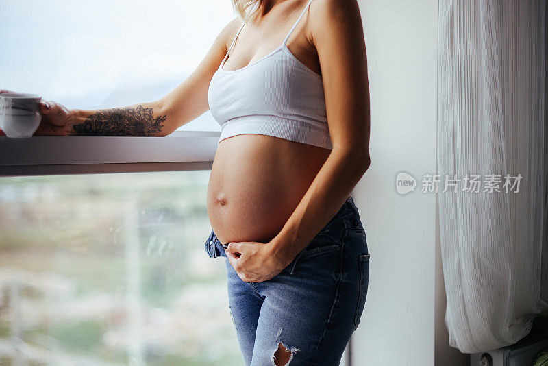 年轻快乐的孕妇站在窗边喝咖啡的肖像