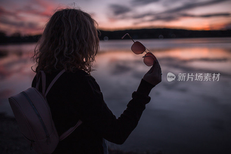 一位面目全非的女子摘下墨镜，在湖岸欣赏日落