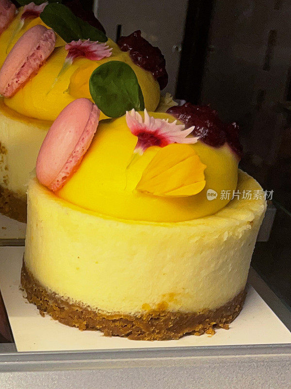 面包店展示的两个黄色芒果芝士蛋糕的特写图像，白色盘子，烘烤和顶部的马卡龙和花瓣，美食甜点，重点在前景