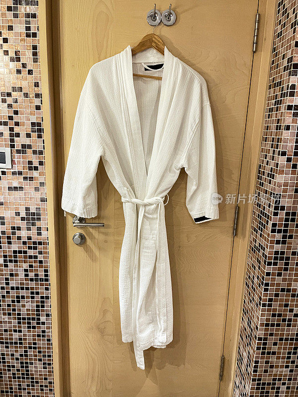 酒店形象，木质衣架上的白色毛巾袍挂在木门背面的铬钩上，酒店浴室，棕色马赛克瓷砖墙，重点在前景