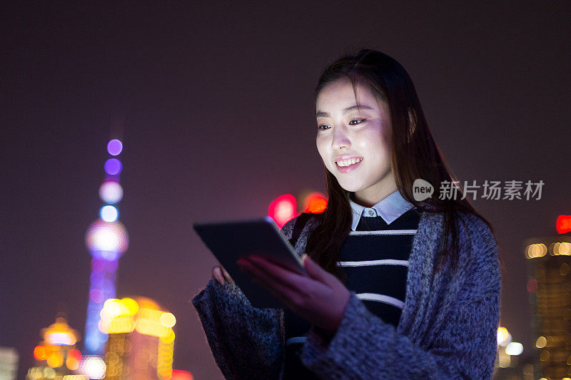 年轻美丽的亚洲女人晚上在现代城市玩数字平板电脑