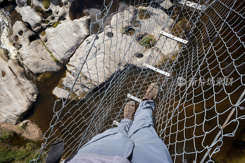 一个穿着灰色裤子的徒步旅行者穿过河上岩石上的吊桥的观点
