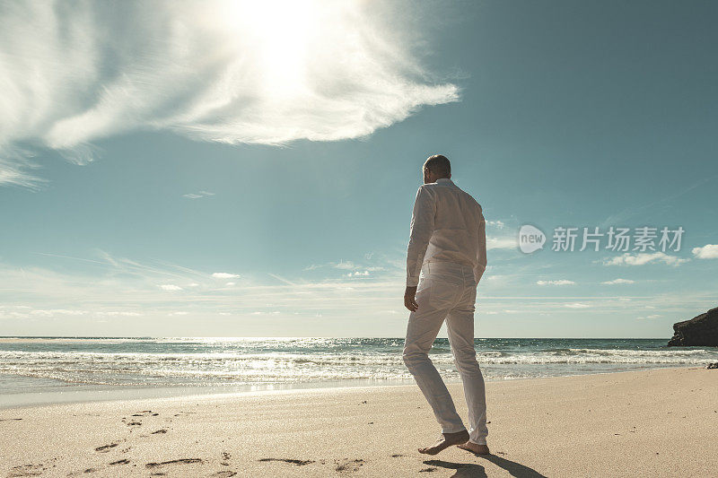 穿着一身白衣服的人在阳光明媚空旷的海滩上走向水边。