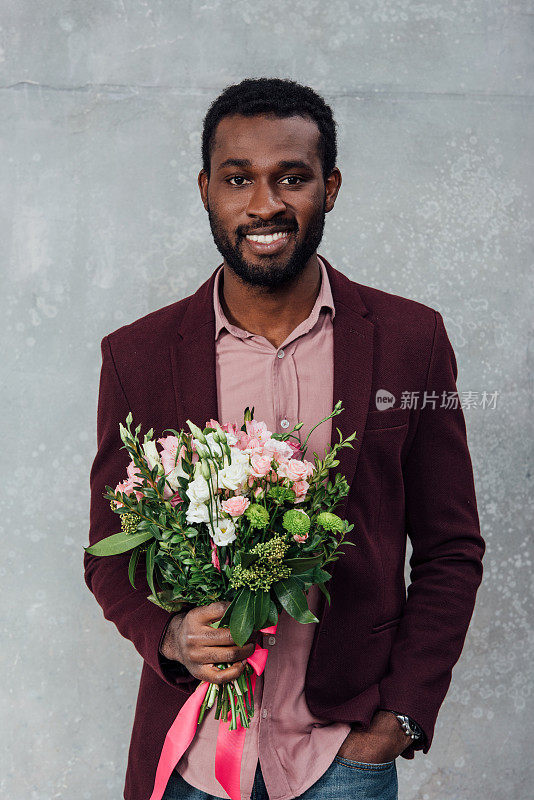 非裔美国人穿着休闲衣服，拿着鲜花，看着灰色背景上的照相机