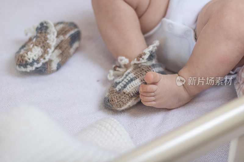 刚出生的婴儿的脚紧裹在羊毛袜子上，躺在白色的毯子上。婴儿在婴儿床里。一只袜子从脚上取下来放在旁边，一只婴儿的脚是光着的。脚冻结