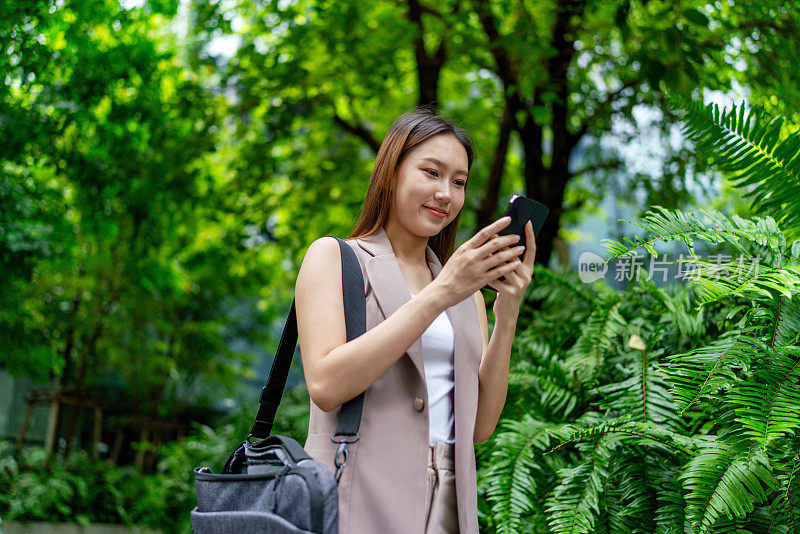 移动中的商业。自然、平衡和互联的移动性。一位年轻的亚洲女商人背着笔记本电脑包，用智能手机上下班，她在中央商务区上班，背景是郁郁葱葱的绿树和花园