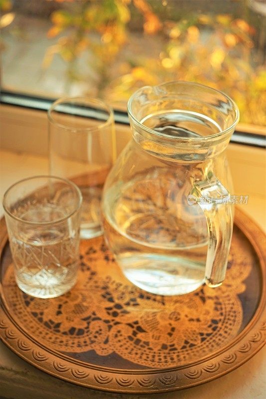 一壶水和一个玻璃杯放在窗台上的一个老式木托盘里。