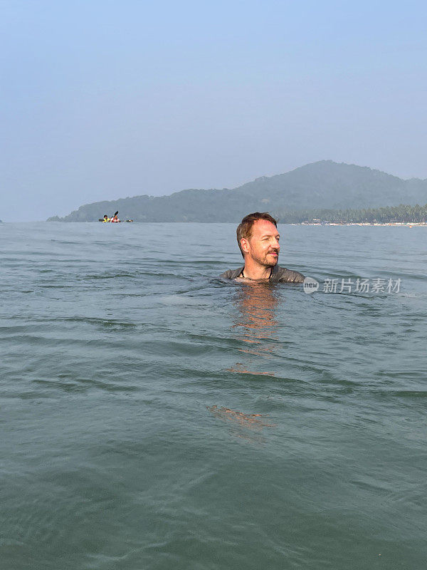 白人男子在海滩度假，穿着衬衫在海里游泳和戏水的形象，水面水平闪闪发光，波涛汹涌的水面波浪，重点放在前景