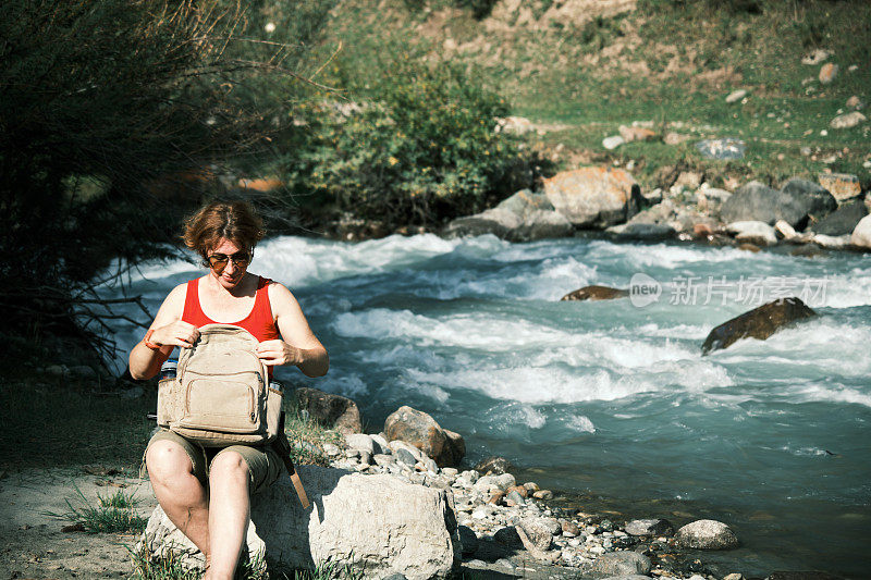 一位女游客坐在山河边，翻看她的背包