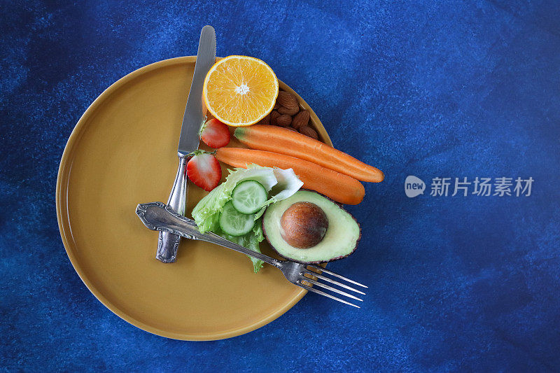 黄色盘子的形象，不锈钢刀叉形成钟面和指针，健康的水果和蔬菜在小段，蓝色背景，高架视图，复制空间，饮食和间歇性禁食的概念