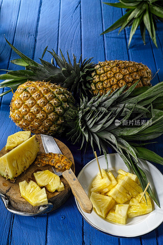 新鲜切好的热带菠萝块放在盘子里，旁边是用刀切好的菠萝，背景是两个新鲜的菠萝。