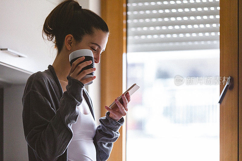 年轻女子在家里，用她的智能手机和喝咖啡。