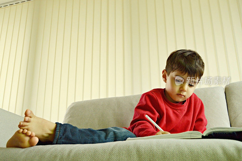一个可爱的学龄前儿童穿着红色的套头衫和蓝色的牛仔裤，在笔记本上用白色的铅笔随意地坐在客厅的白色沙发上写东西