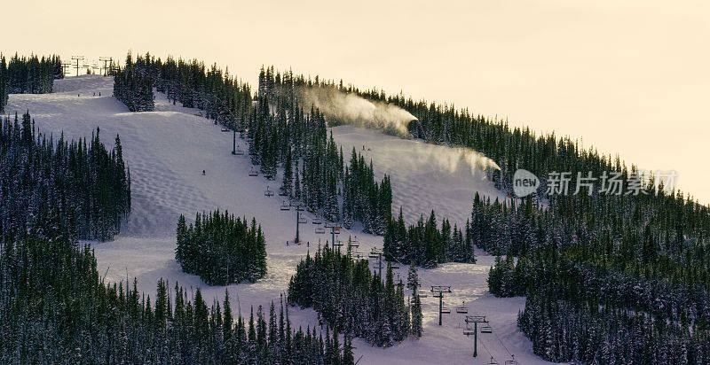 冬季日落时分，在科罗拉多山滑雪场，当人们滑雪或滑雪时，雪机将雪吹向空中(比弗溪)