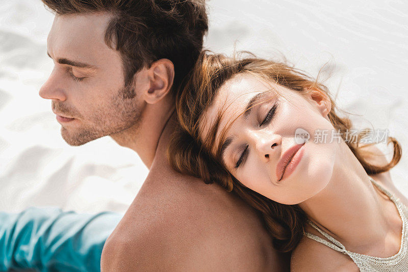 微笑的年轻夫妇背靠背坐在海滩上