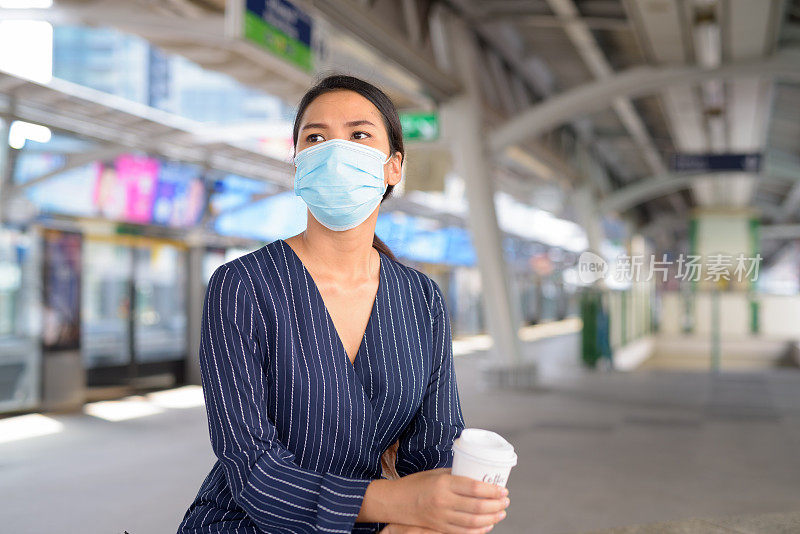 年轻的亚洲女商人戴着面具，一边喝着咖啡，一边在轻轨站等待