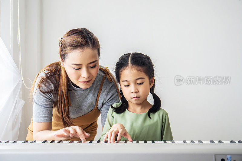 快乐的小亚洲女儿弹钢琴与母亲在家里，母亲教女儿弹钢琴，他们弹琴唱歌。他们玩得很开心。
