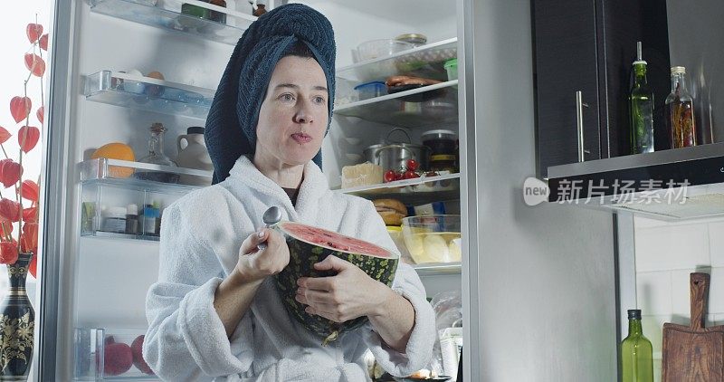 成熟的女人穿着浴袍和头巾在冰箱旁边吃着半个西瓜