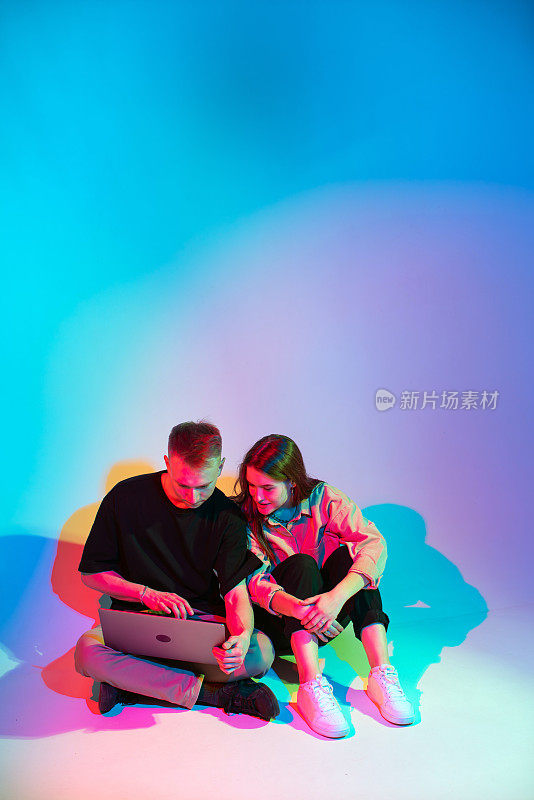 酷时尚的年轻夫妇与笔记本电脑。彩色的霓虹灯。女孩和男孩的博客为社交网络创造内容