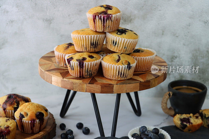 近景:木头和金属蛋糕架堆放着自制的蓝莓松饼，放在纸制蛋糕盒里，旁边是灰色的平纹棉布和咖啡杯，周围是蓝莓，大理石效果的背景，聚焦前景