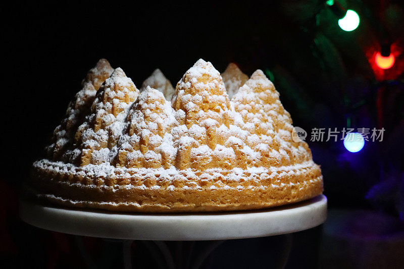 自制的特写图像，圣诞树形状，白色的柠檬毛绒绒的Bundt蛋糕，大理石和金属蛋糕架，柠檬味的环海绵覆盖着糖霜，照亮的精灵灯，黑色背景，聚焦前景