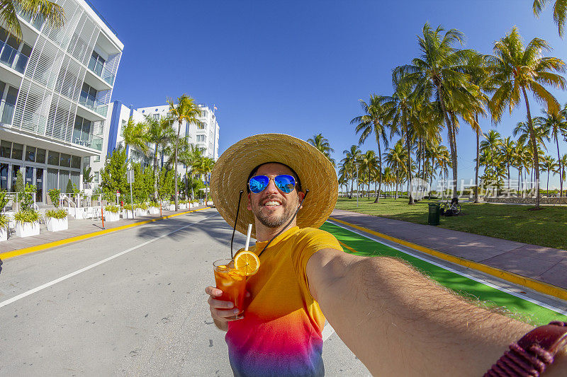 美国南佛罗里达迈阿密南海滩，一名年轻的旅行男性在海洋大道前享受他的假期，一边喝着鸡尾酒，视角自拍