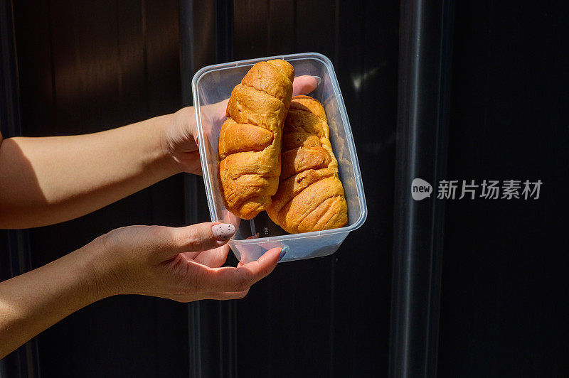 女人的手拿着一个塑料盒子与圆面包和牛角面包与铁金属背景