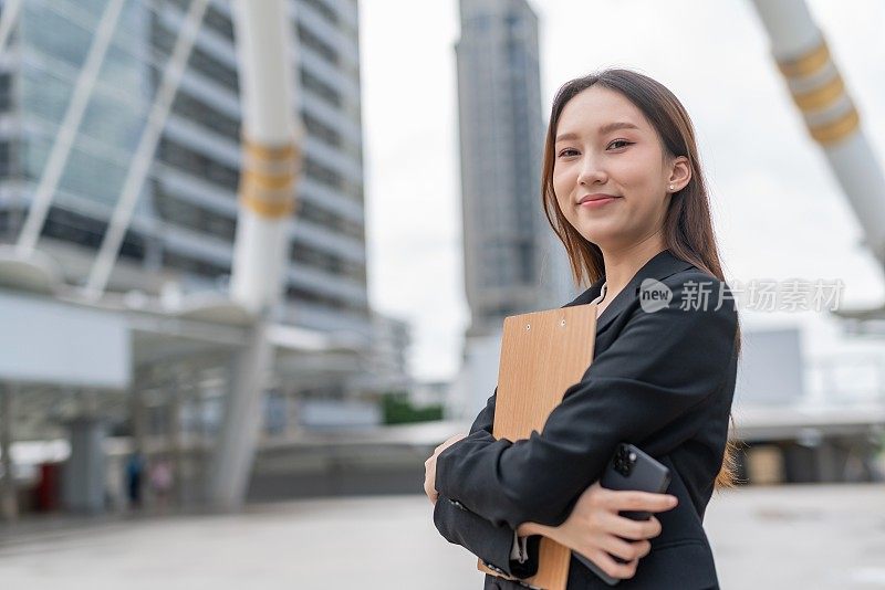 迷人的年轻亚洲职业女商人女士，带着美好的自信的微笑，看着相机拿着夹夹板和智能手机在曼谷市的中央商业区