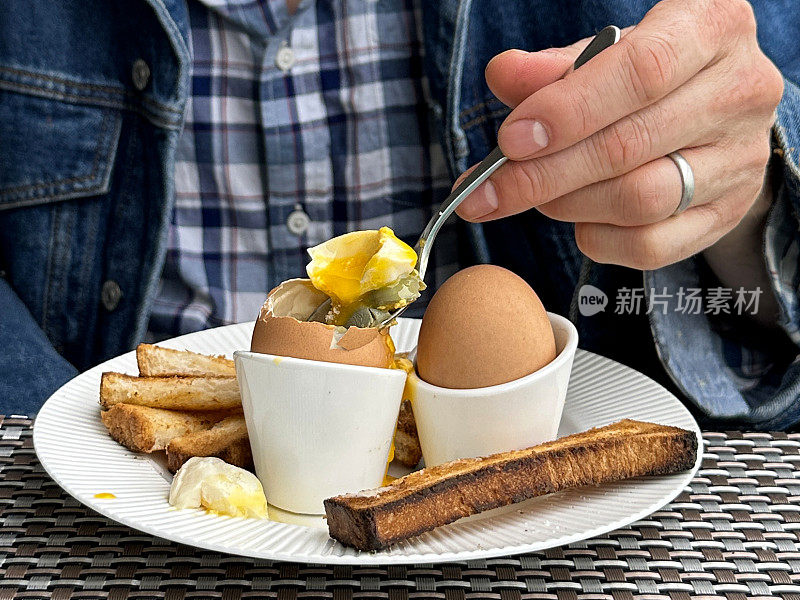 全画幅图片，白色的盘子里有黄油吐司士兵，两个溏心蛋放在蛋杯里，上面有溏心蛋和溏心蛋黄，被一个不认识的人用茶匙吃着，重点在前景