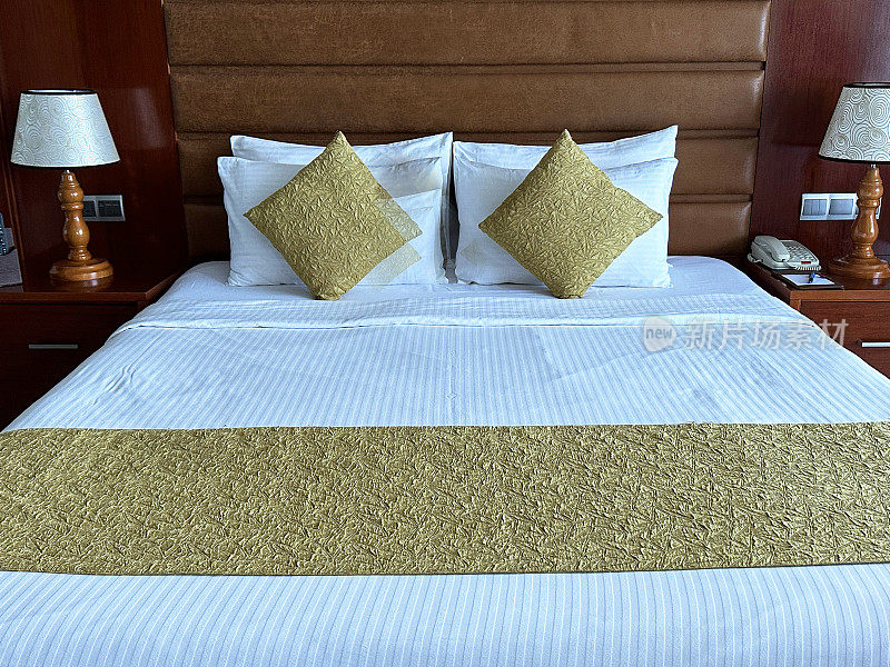 酒店房间双人床的全画幅图像，白色酒店枕头倚在人造革床头板上，白色羽绒被，金色靠垫，床头柜上的电灯，重点在前景