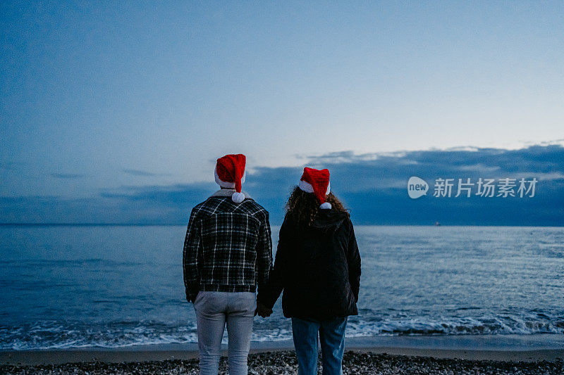希腊，一对戴着圣诞帽的年轻夫妇在黎明时分欣赏着海岸线的景色