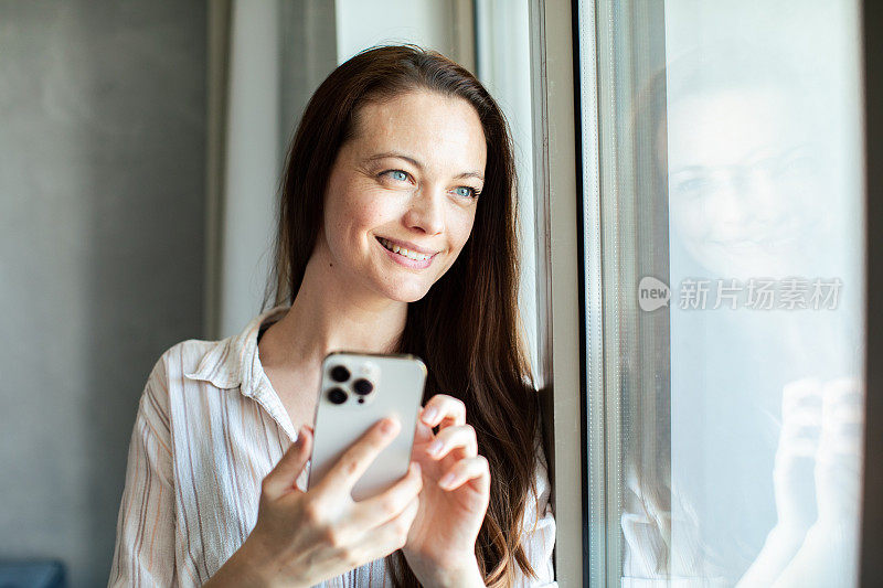 年轻女子在家里一边看窗外一边用智能手机