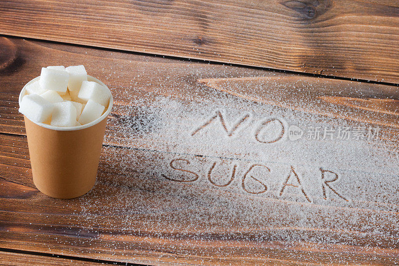在棕色的背景上，有一个装满精制糖的纸玻璃杯，一堆糖上写着“不加糖”的字样。减少摄入和放弃糖的概念