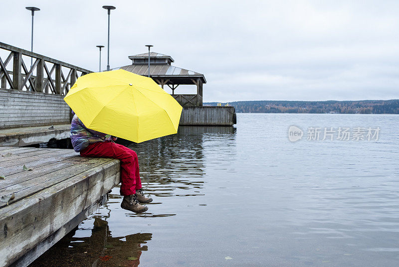 一个8岁的小女孩撑着一把大黄伞坐在木码头上，欣赏着秋天的景色。