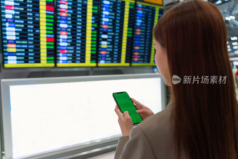 后视图:年轻的亚洲Z世代女性手持手提包，手持智能手机，站在机场候机楼的FIDS登机牌前。出差的商务人士。手持智能手机的年轻亚洲女性旅客在机场候机楼。