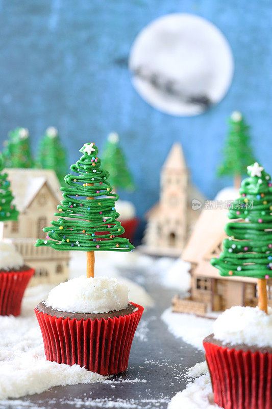 圣诞村场景特写图片，批量自制，圣诞树设计巧克力纸杯蛋糕，模型木屋，糖霜雪，圣诞老人雪橇和驯鹿剪影飞过月亮的背景，重点在前景