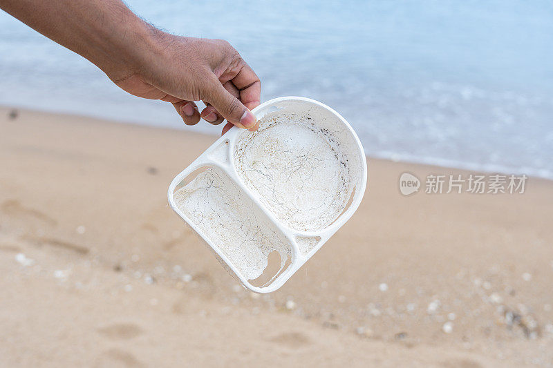 近距离拍摄的人的手捡塑料食品容器垃圾在海滩上清理凌乱和肮脏的海滩。一个不知名的志愿者保护环境免受塑料垃圾污染的破坏。