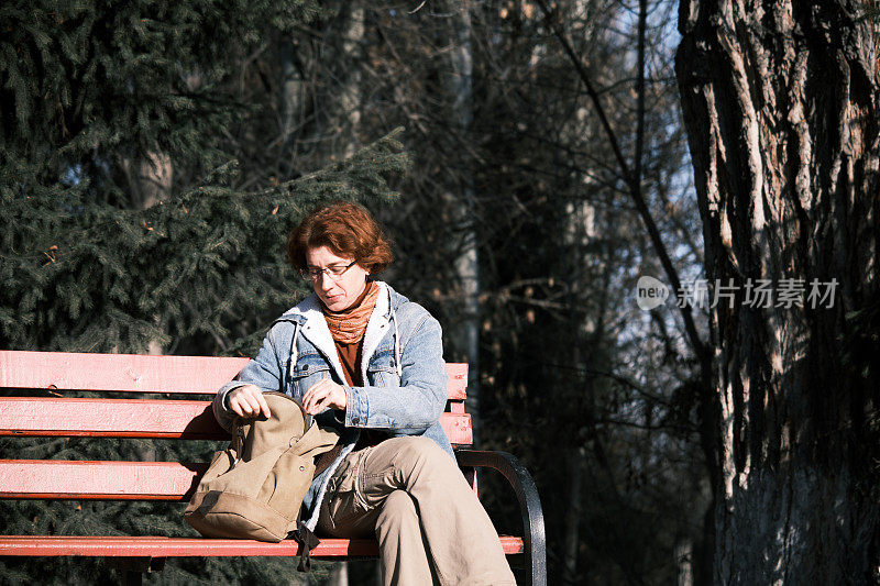 单身的成熟女人坐在公园的长椅上，在背包里找东西