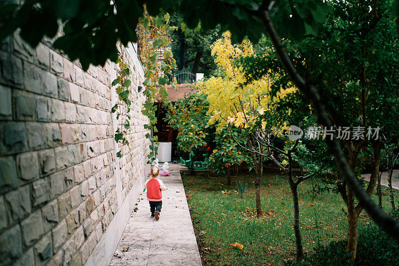 小女孩沿着花园的一条小路沿着石栅栏走着。后视图
