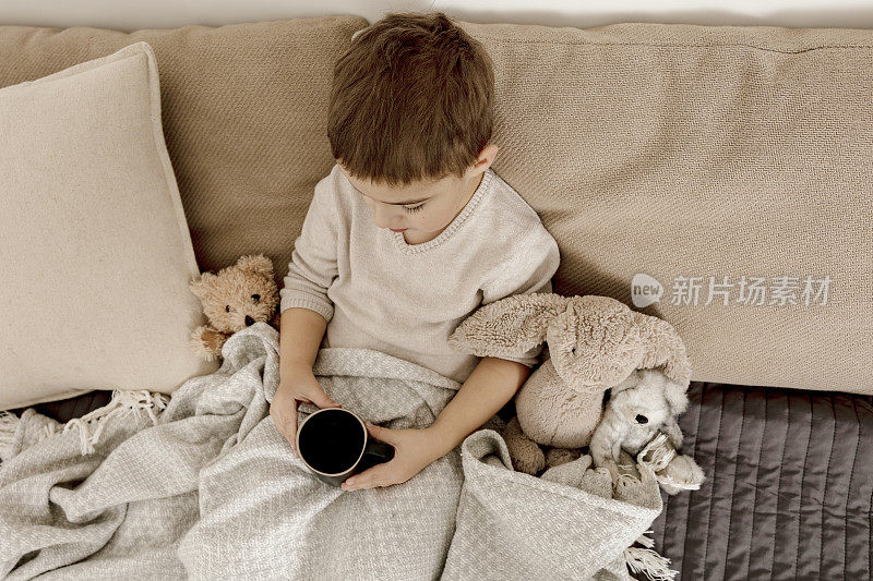 生病的小男孩在家里的床上喝热茶。生病的孩子用毯子裹着，房间里放着马克杯。流感季节。室内装饰和衣服采用自然的大地色。舒适的环境。