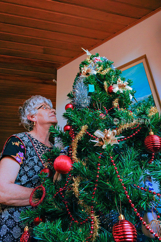 一位白发苍发的拉丁美洲高级女士正在为她公寓客厅里的一棵人造圣诞树做最后的装饰，非常专注地为家人装饰。