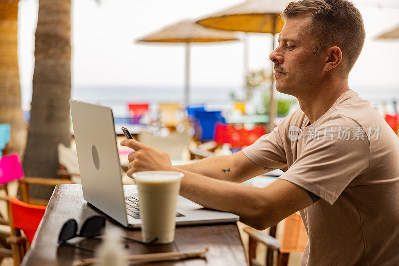 在海滩咖啡店里，男人一边用笔记本电脑工作，一边用手机