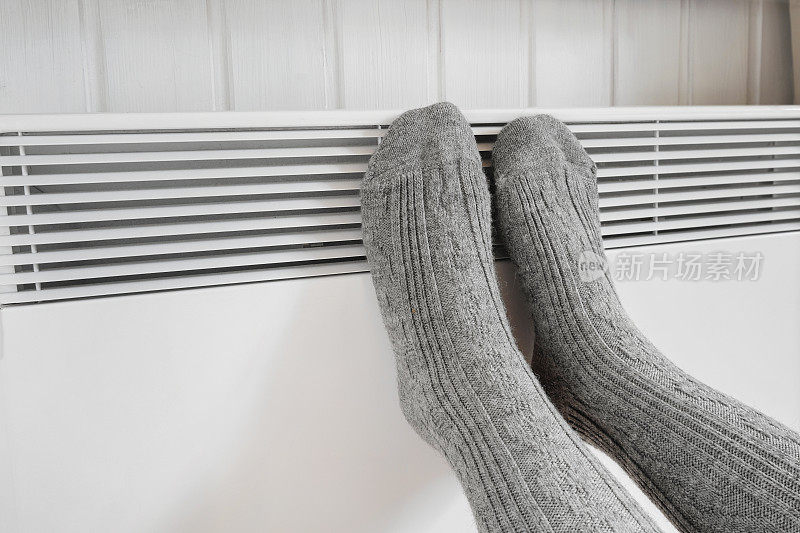 穿着针织袜的腿在电加热器上加热。冰冻的女人穿着温暖的羊毛袜子冻在冬天的寒冷。不舒服的呆在家里。女孩在现代暖气片上暖脚