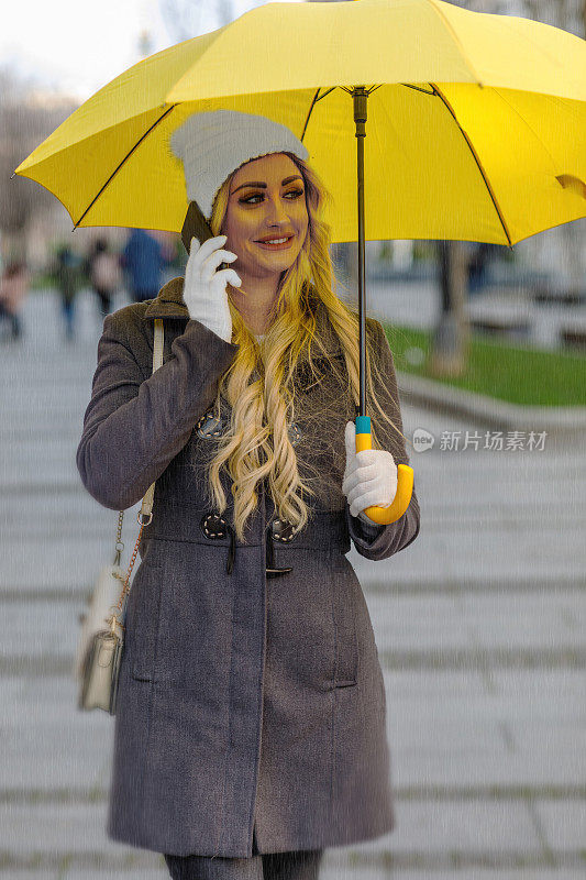 一个迷人的年轻女子在雨天走在城市街道上使用手机的肖像。