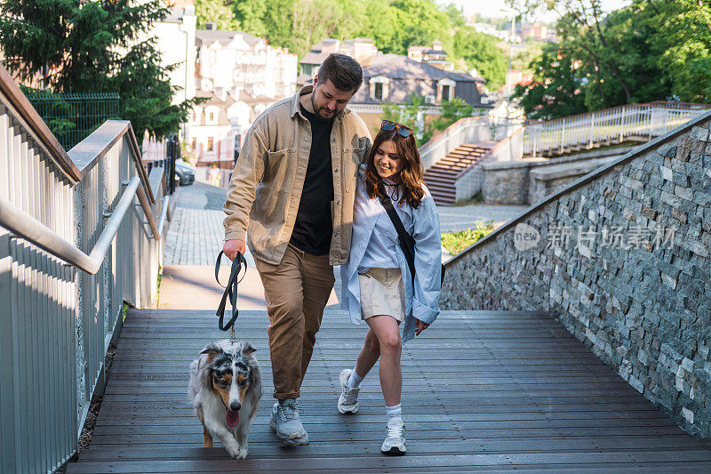 一对年轻夫妇和他们的澳洲牧羊犬在市区散步。幸福的男人和女人在城市里遛宠物，真正的生活方式