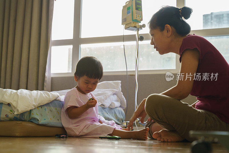 生病的小男孩坐在地板上，在医院病房的恢复与母亲的安慰和照顾他。可爱的亚洲孩子感染病毒住院了。婴儿保健医疗理念。
