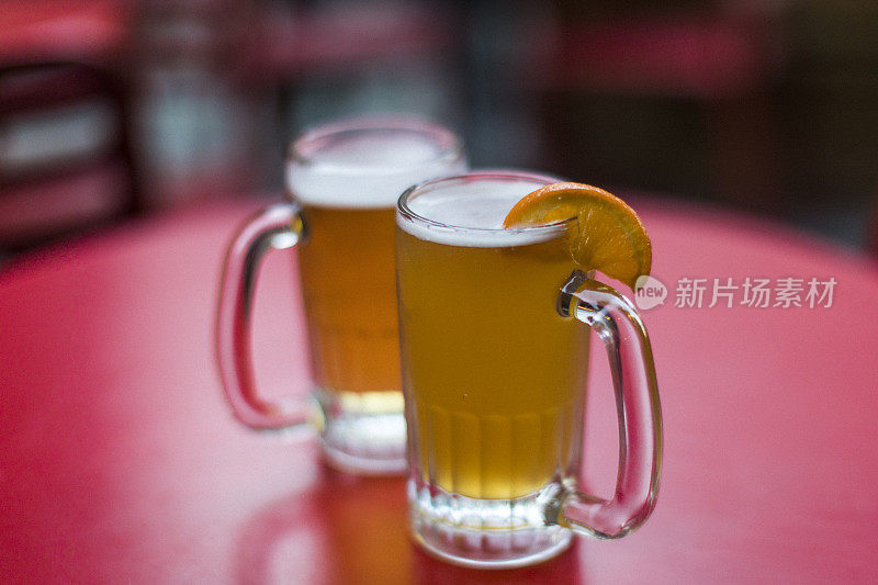 两个清爽的啤酒杯放在酒吧露台的红桌上，一个是杏子啤酒，另一个是奶油啤酒。