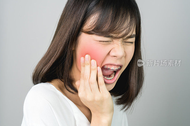 亚洲女孩穿着白色休闲装炫耀牙痛，也许是因为没有保持良好的口腔健康。在灰色的背景下会发出柔和的光。