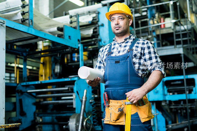 认真的混合种族工业工程师在整体和硬帽工作在工厂:他拿着轧制蓝图和行走在制造工厂